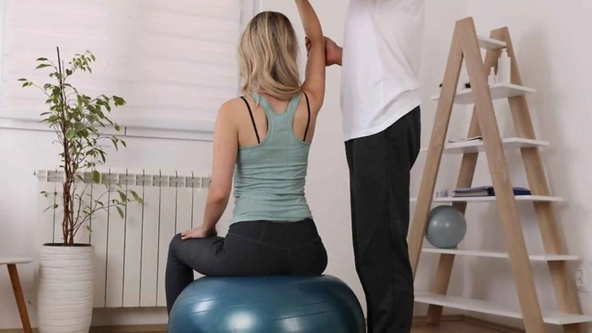 A lady with her physiotherapist on a gym ball with her arm raised